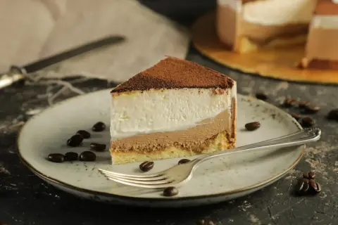 Pyszny sernik z mascarpone, posypany kakao, na talerzu z ziarnami kawy. Obok widelca leży kawałek ciasta.