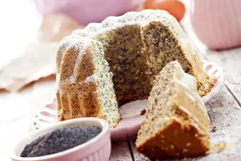 Pyszna babka makowa, oprószona cukrem pudrem, z widocznym ziarnem maku. Obok miseczka z makiem i kawałek ciasta.