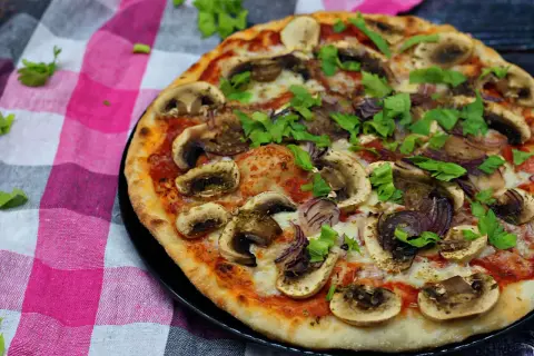 Pyszna pizza z pieczarkami na pizze, cebulą i świeżą natką pietruszki, podana na kraciastej serwetce.