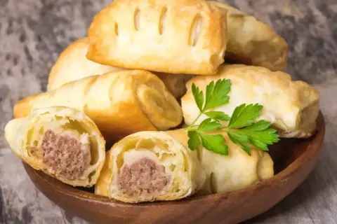 Stos pieczonego ciasta francuskiego z kiełbasą w środku, ozdobione natką pietruszki. Idealne na szybką przekąskę.