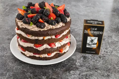 Wielowarstwowy tort czekoladowy z kremem mascarpone, udekorowany świeżymi truskawkami, jeżynami i miętą. Obok stoi opakowanie kakao DecoMorreno.