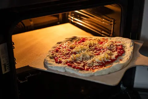 Pizza z sosem i serem na desce, gotowa do włożenia na rozgrzany kamień szamotowy w piekarniku.