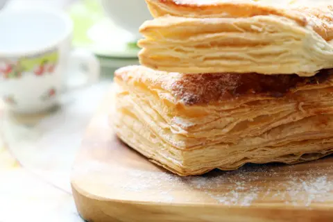 Stos puszystego ciasta francuskiego, idealnego do nauki, jak zrobić ciasto francuskie. Posypane cukrem pudrem, na drewnianej desce.