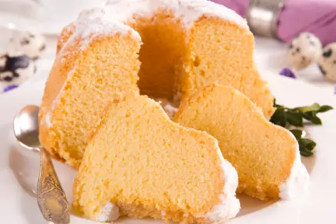 Puszysta babka z mascarpone, oprószona cukrem pudrem, z kilkoma kawałkami na talerzu. Obok leży łyżeczka.