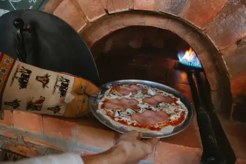 Ręka w rękawicy z krowami wkłada pizzę do pieca szamotowego. Płomień tańczy w głębi pieca.