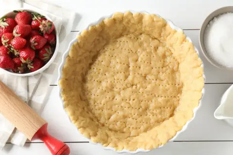 Gotowe ciasto na tartę z truskawkami czeka na nadzienie. Obok miska świeżych truskawek, cukier i dzbanuszek mleka.