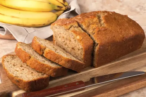 Pyszna babka bananowa, pokrojona na kromki, leży na desce obok pęczka bananów.