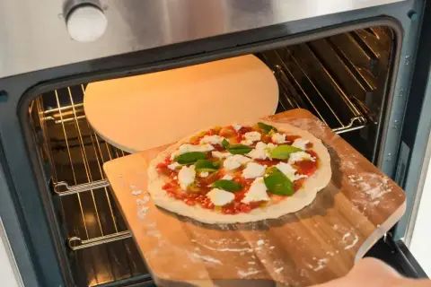 Przygotowania do dnia pizzy 2026: świeża pizza z mozzarellą i bazylią wsuwana na rozgrzany kamień w piekarniku.