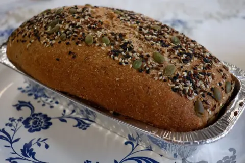 Chleb Ezechiela w formie bochenka, posypany pestkami dyni, sezamem i siemieniem lnianym, w aluminiowej foremce.