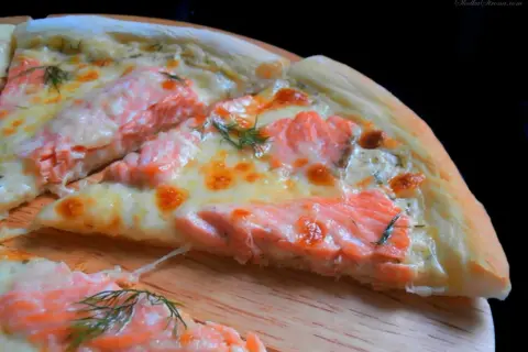 Kawałek pysznej pizzy z łososiem, roztopionym serem i koperkiem na drewnianej desce.