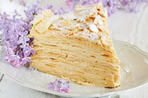 Pyszny tort napoleon z delikatnym kremem i chrupiącymi warstwami ciasta, ozdobiony płatkami migdałów i cukrem pudrem, w otoczeniu kwitnącego bzu.
