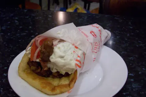 Pyszna pita grecka z mięsem, warzywami i sosami, zawinięta w papier z greckimi napisami.