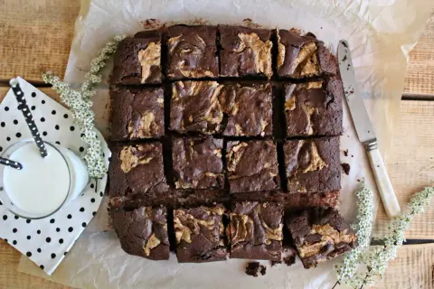 Pyszne brownie z masłem orzechowym, pokrojone w kostkę, czeka na degustację. Obok szklanka mleka i delikatne białe kwiaty.