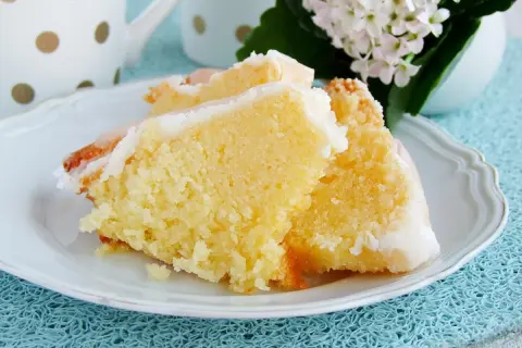 Puszysta babka jogurtowa z lukrem na białym talerzyku, obok filiżanka i kwiaty.