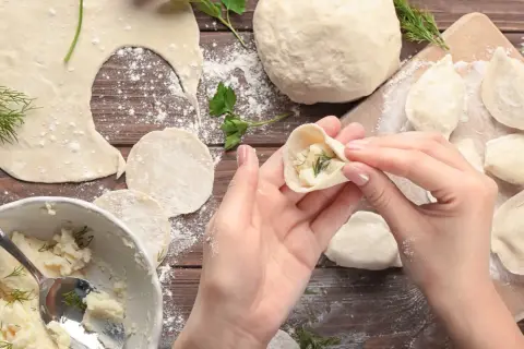 Ręce formują pieroga z ciasta na pierogi, wypełnionego farszem ziemniaczanym i koperkiem. Na drewnianym blacie widać mąkę, gotowe pierogi i miskę z farszem.