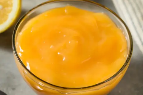 Gładki, żółty lemon curd w szklanej miseczce, obok połówka cytryny. Idealny do deserów.