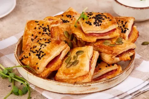 Pizzerinki z ciasta francuskiego, posypane czarnuszką i pestkami dyni, ułożone na talerzu. Idealne na przekąskę.