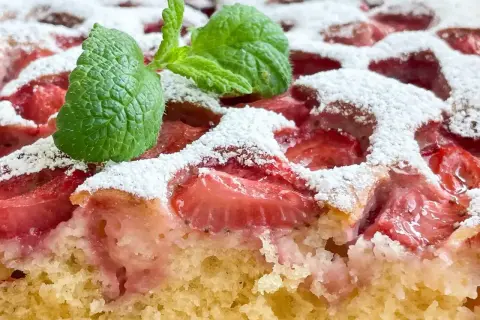Puszyste ciasto jogurtowe z truskawkami, posypane cukrem pudrem i ozdobione listkiem mięty. Idealne na letni deser.