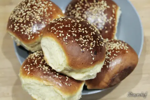 Pięć puszystych, złocistych bułek do burgerów, posypanych sezamem, ułożonych na talerzu.