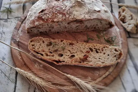 Chleb z suszonymi pomidorami, pokrojony na desce, z gałązką rozmarynu i kłosami zbóż.