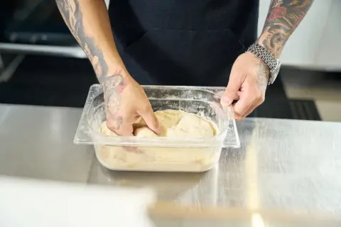 Ręce z tatuażami mieszają ciasto na pizzę w pojemniku. Poznaj prosty sposób, jak rozmrozić ciasto na pizzę, by było idealne.